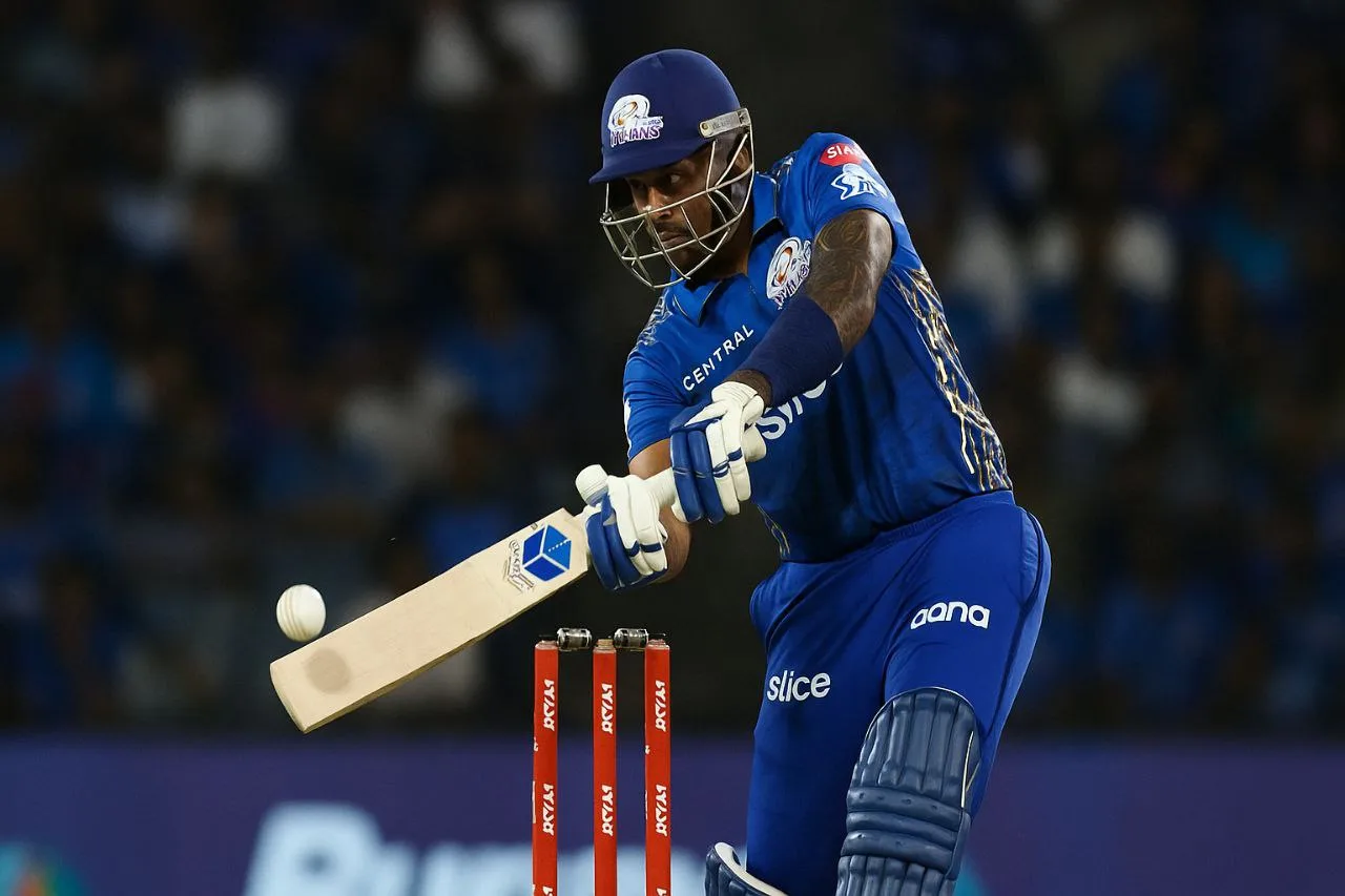 Suryakumar Yadav plays a scoop shot wearing the Mumbai Indians IPL jersey during a night match, with the stadium crowd blurred in the background.
